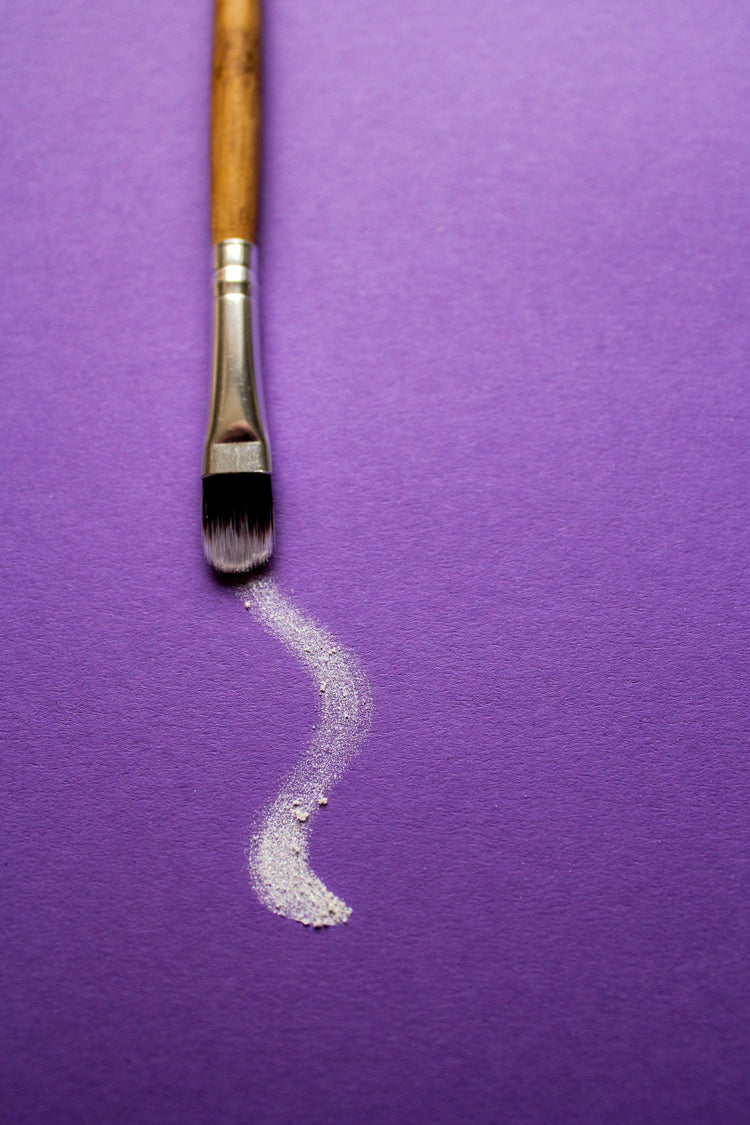 A paintbrush rests on purple paper, a long white curl of dusty white paint traces a path | Photo by: Aelis Harris