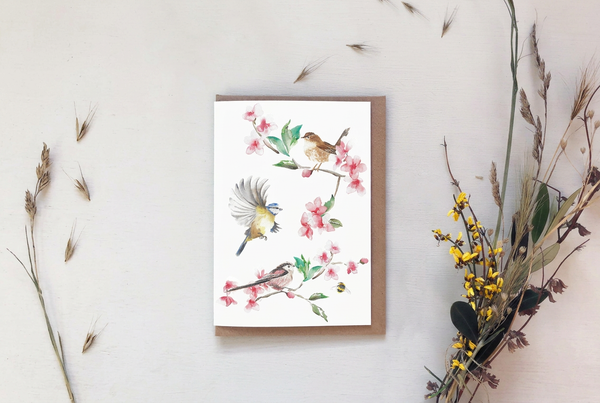 A vertical art print featuring watercolor birds perched on pink flowering branches, displayed on a kraft envelope against a light neutral background with dried floral props.