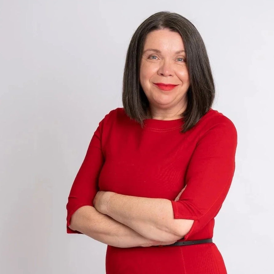 Author Claire_Gelder, with shoulder-length dark hair stands against a light grey studio background, wearing a bright red fitted dress with three-quarter sleeves and a thin dark waistband, with her arms crossed.