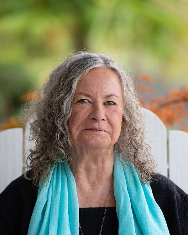 A portrait of Author Deb Bowen, with long, curly gray hair wearing a bright turquoise blue scarf and a black top, seated outdoors in front of a white chair with a blurred green garden background.