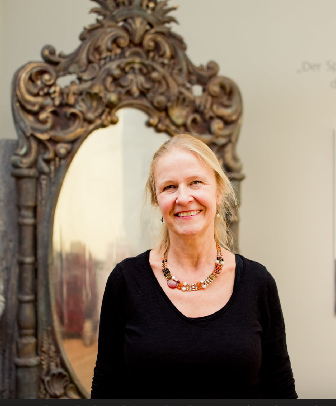 Woman standing in front of an ornate mirror, wearing a black top and a colorful necklace.