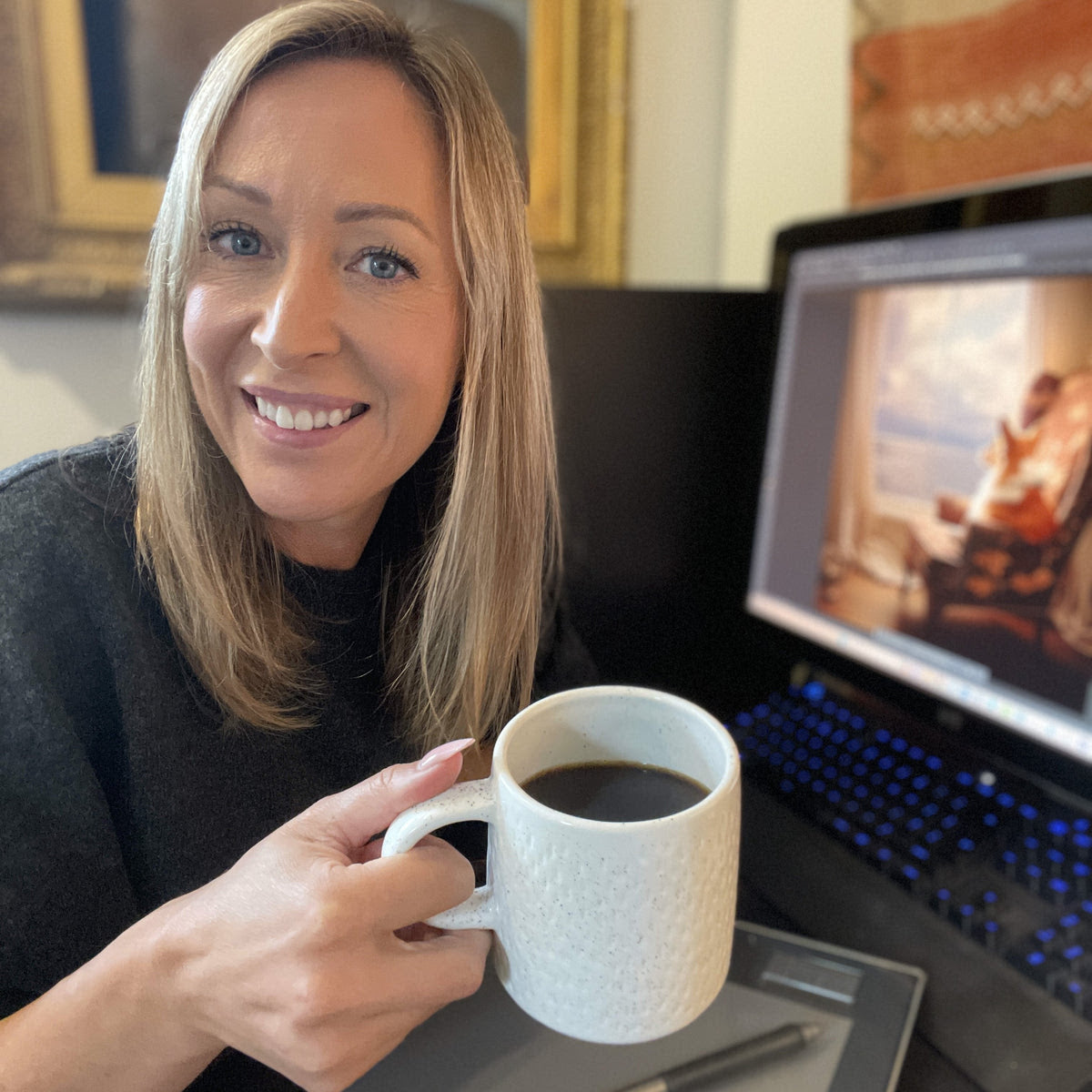 Author Victoria Beata smiling , holding a mug of hot coffee , in front of her computer screen showing her art illustration   