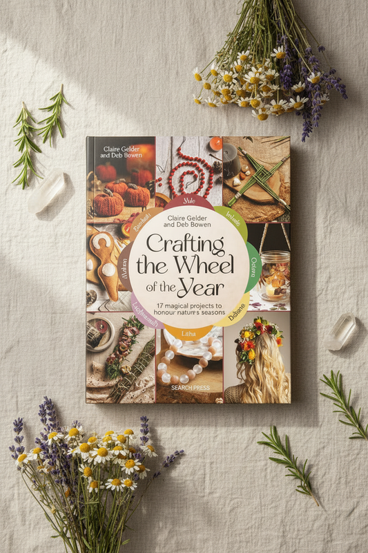 A book titled 'Crafting the Wheel of the Year' by Claire Goldner and Deb Bowen is displayed on a textured cream linen background, surrounded by dried flowers, green sprigs, and small crystals. The book cover features a collage of seasonal crafting and food imagery with a central white circular title panel.