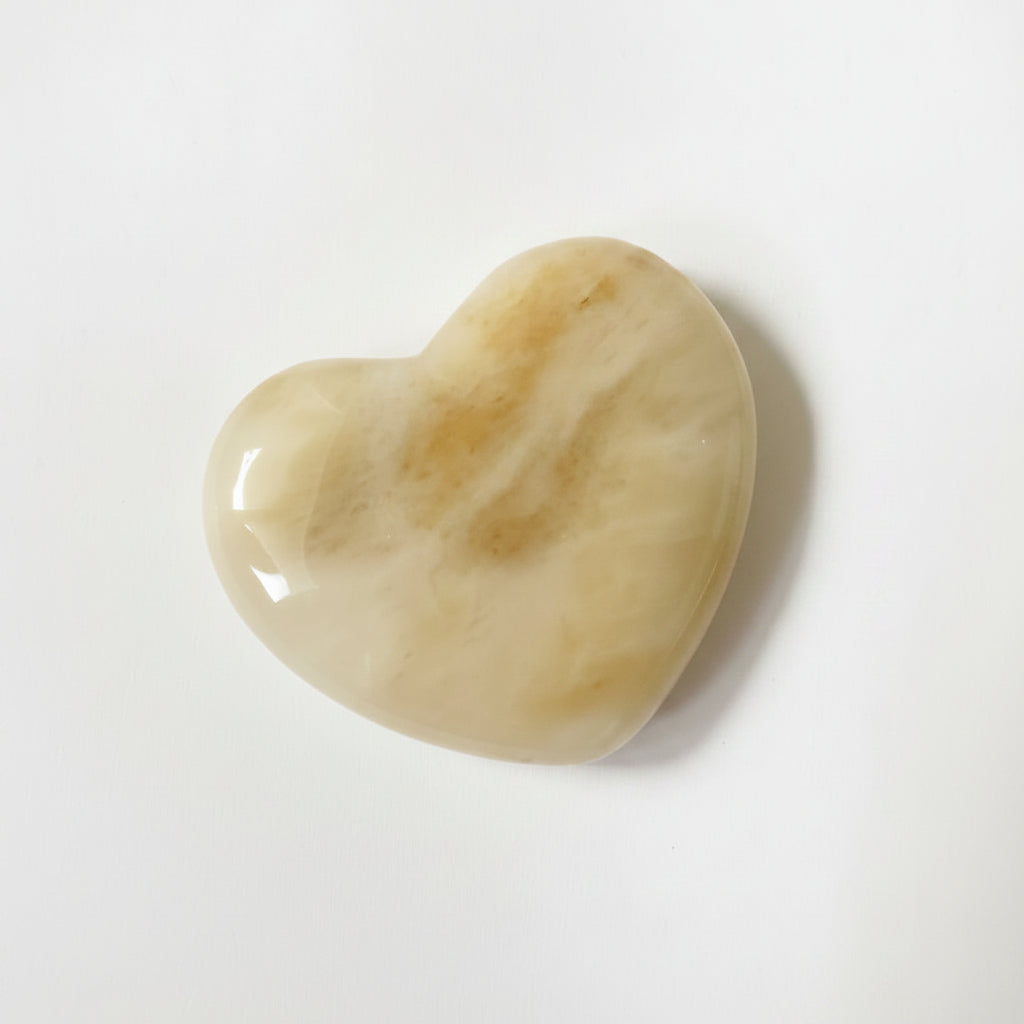 A polished heart-shaped stone with a smooth, glossy finish, featuring natural marbled patterns in cream, beige, and honey, resting on a plain white surface.