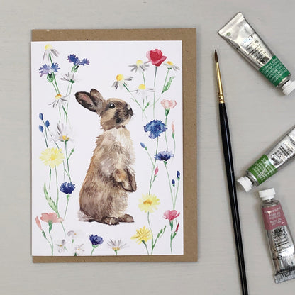 Greeting card with a rabbit and flowers design, accompanied by paint tubes and a brush on a gray background.