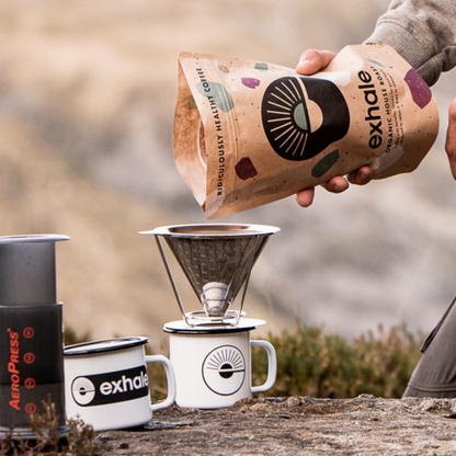 Person pouring coffee from an Exhale Organic Roasters package into a coffee filter over a mug outdoors.
