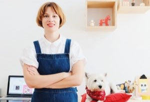 Ekaterina in a blue apron standing in a room with a dog on a red pillow.
