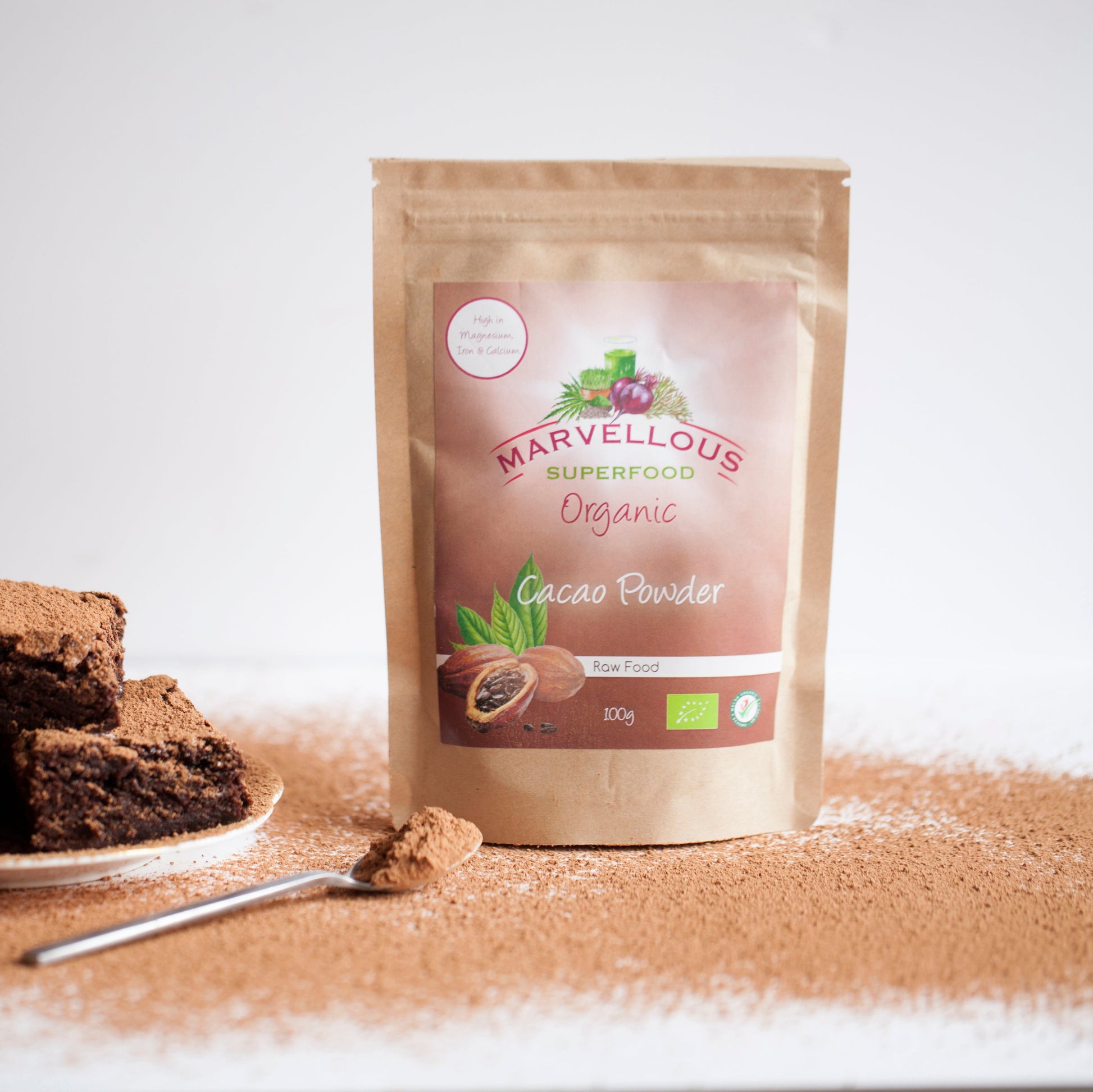 Bag of Marvellous Superfood Organic Cacao Powder with a spoonful of powder and brownies on a white background