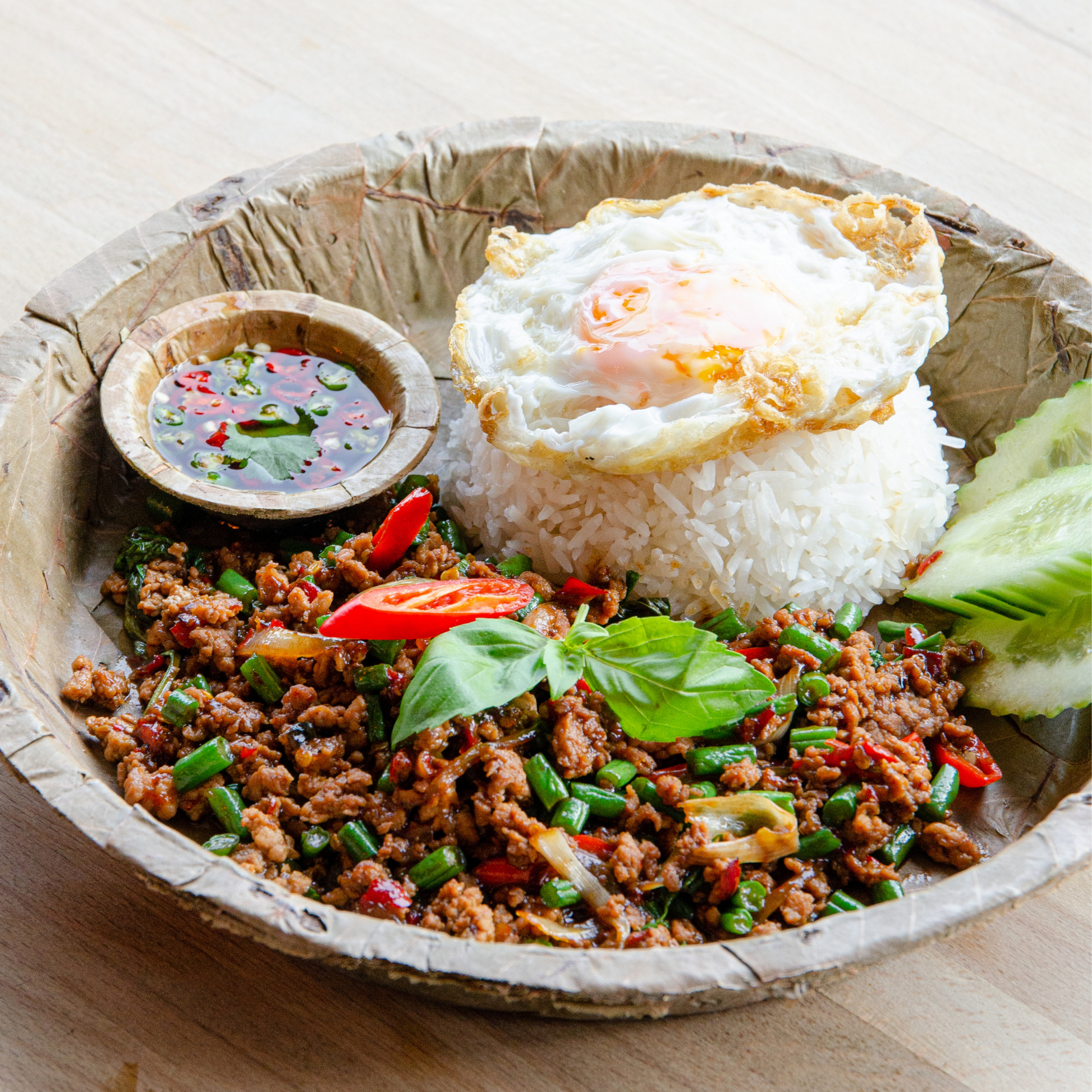 Plated leaf dish with rice, ground meat, vegetables, and a fried egg on a wooden surface.