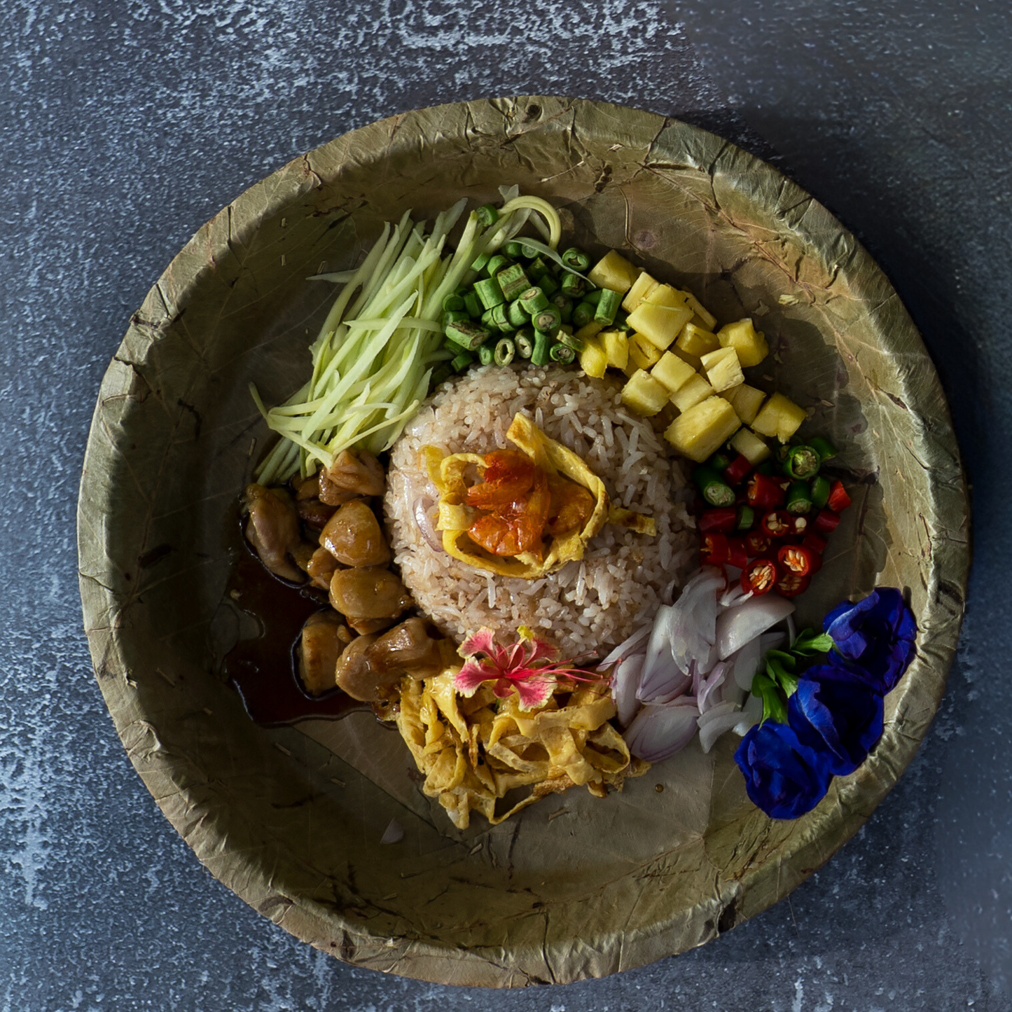 Plated leaf dish with rice, vegetables, and flowers on a textured surface