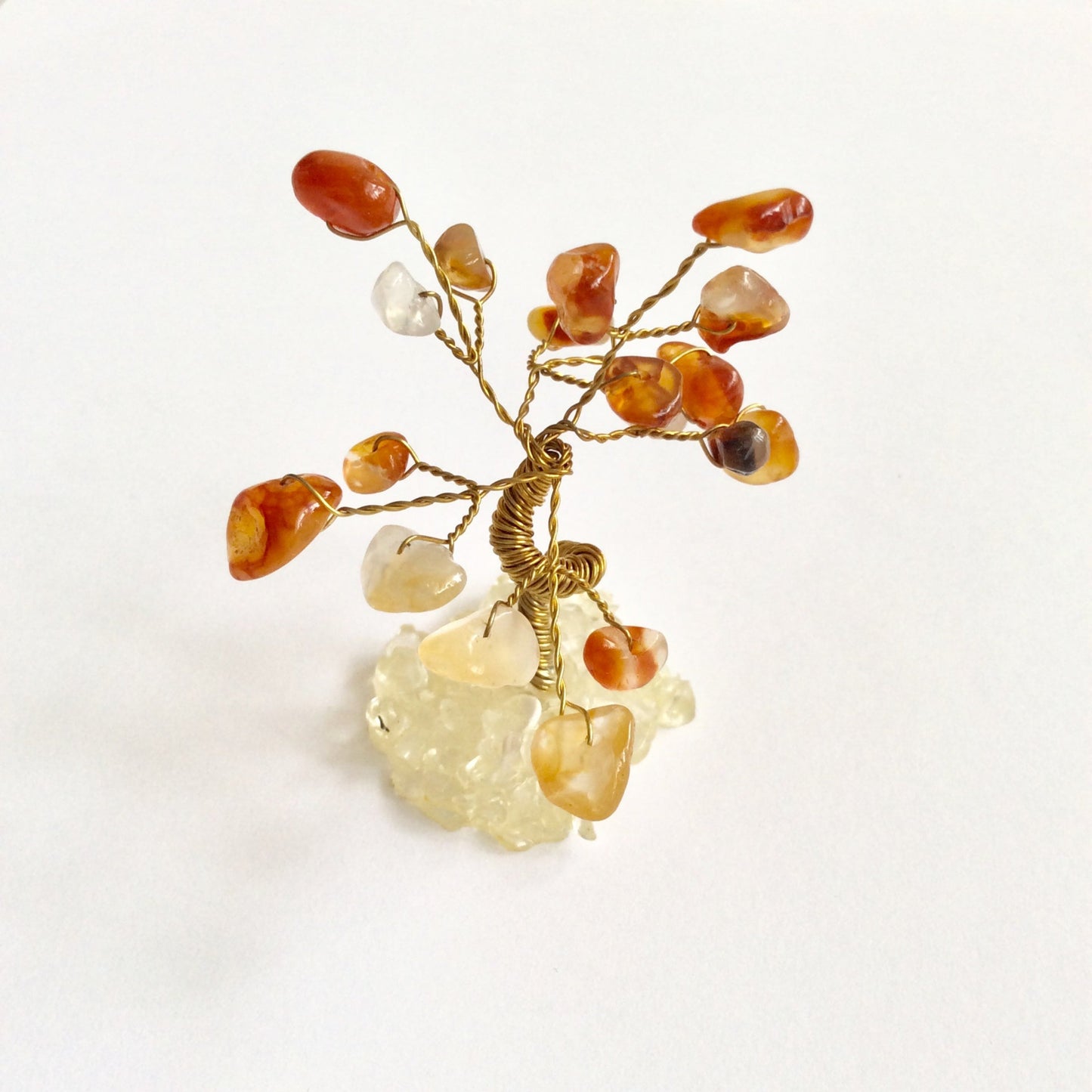 A small decorative wire tree with gold-toned branches and amber, orange, and translucent Carnelian tumbled stones, mounted on a pale crystal base against a plain white background.