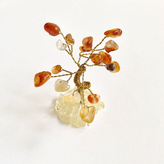 A small decorative wire tree with gold-toned branches and amber, orange, and translucent Carnelian tumbled stones, mounted on a pale crystal base against a plain white background.